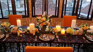 A table set for a meal with candles, flowers, and a view of buildings through large windows. - Hotel Sriti (Magelang)
