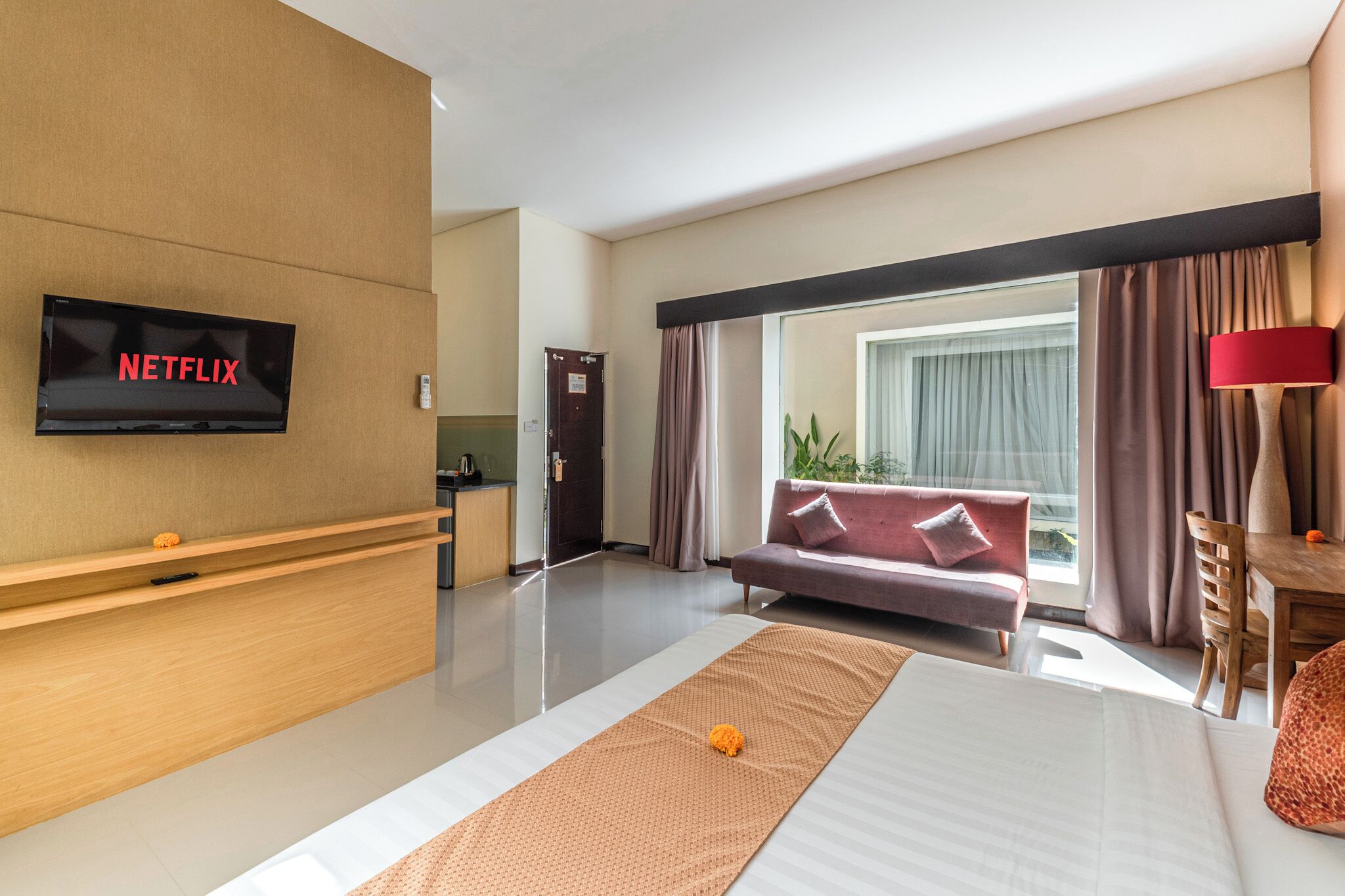 A modern hotel room with a flat-screen TV displaying 'NETFLIX', a sofa, a wooden desk, a dining table, and a large window with curtains.