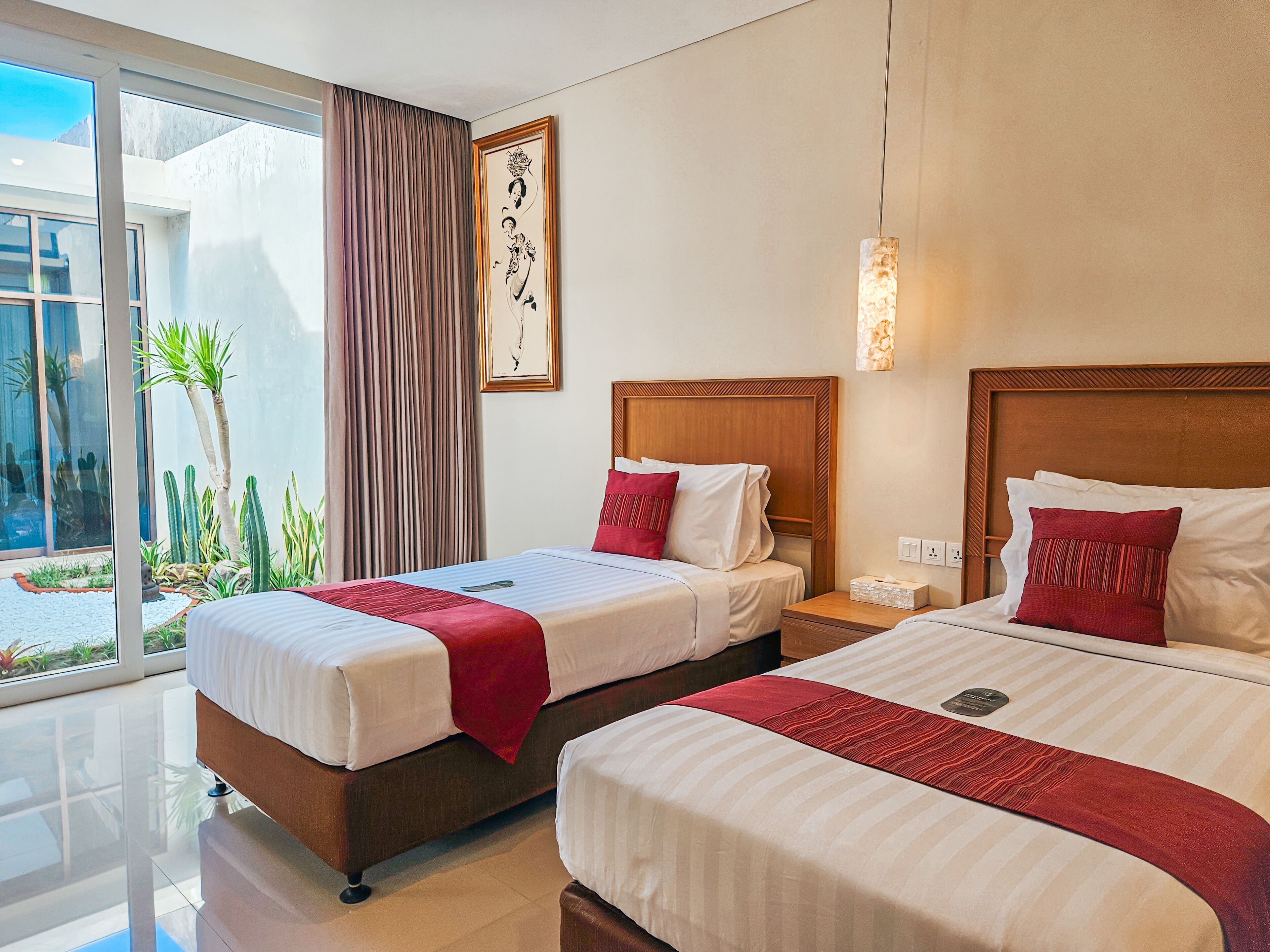A hotel room with two beds, a large window with a view of a building and plants, and a wooden headboard.