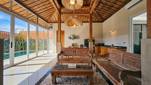 A spacious living area with a high ceiling, wooden beams, a large window, and a patterned floor. - Svarna Suite Seminyak (Legian)