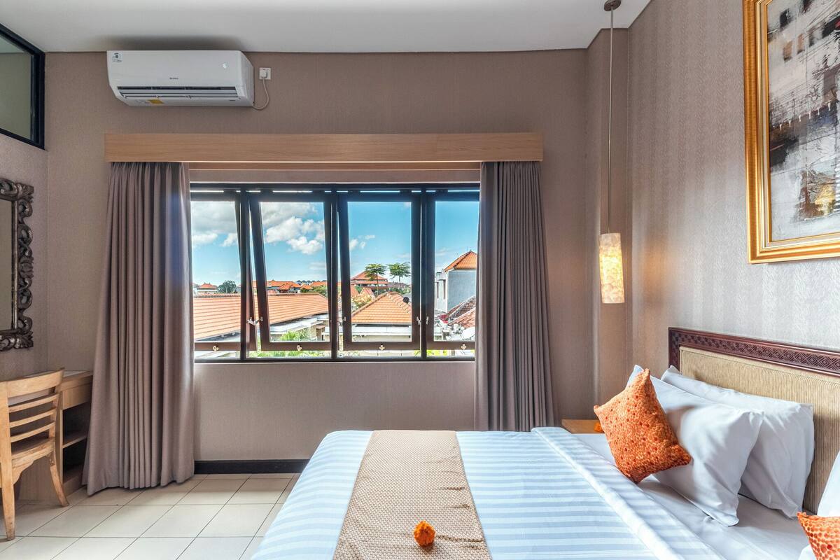 A hotel room with a large window, a bed with pillows, a desk, and an air conditioning unit.