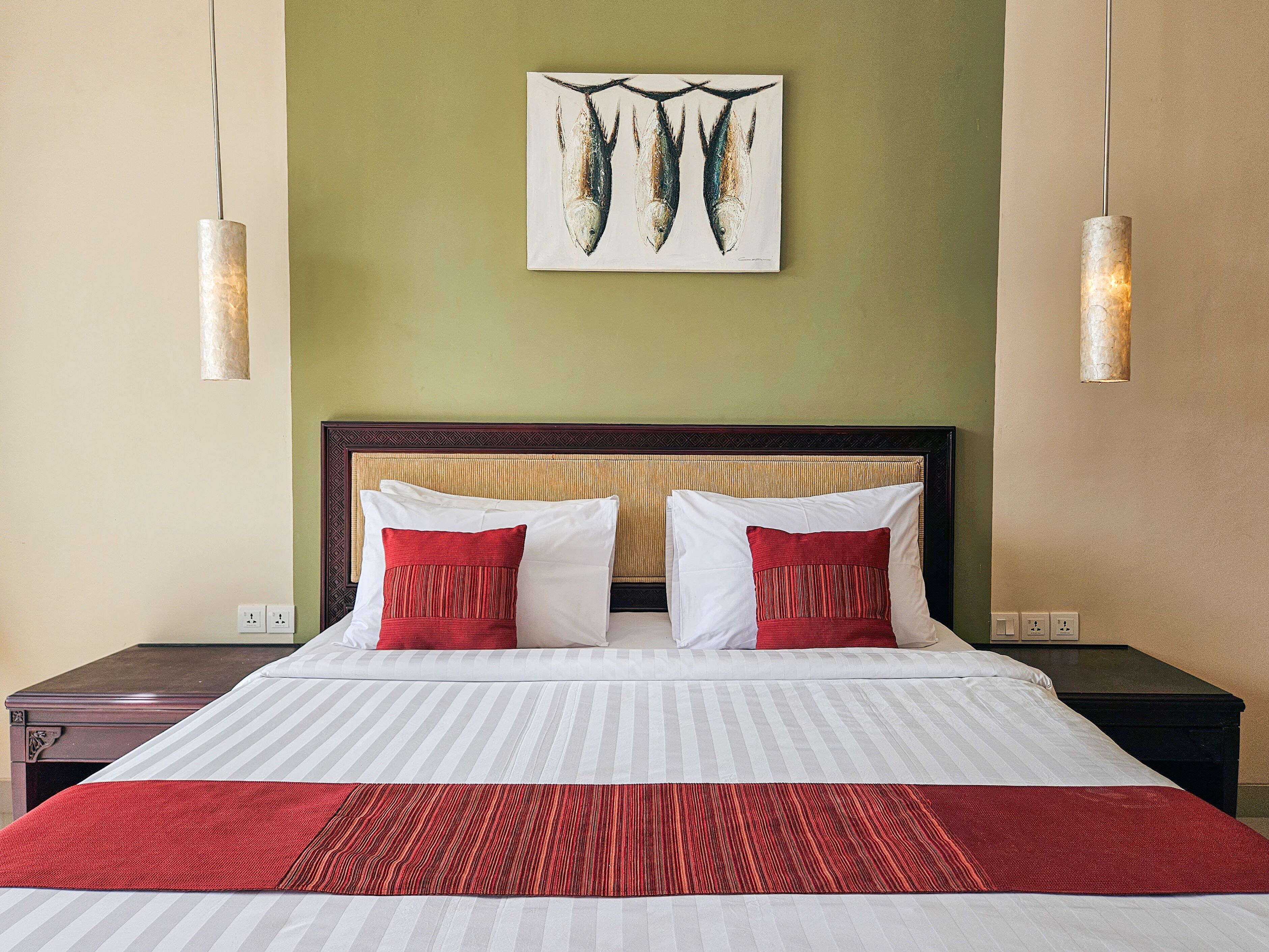Une chambre d’hôtel comprenant un lit, deux tables de chevet, une petite table, une couvre-lit à rayures rouges et blanches, deux oreillers rouges, une couverture à rayures rouges et blanches, une tête de lit en bois, une œuvre d’art représentant des poissons accrochée au mur, et deux lampes suspendues.