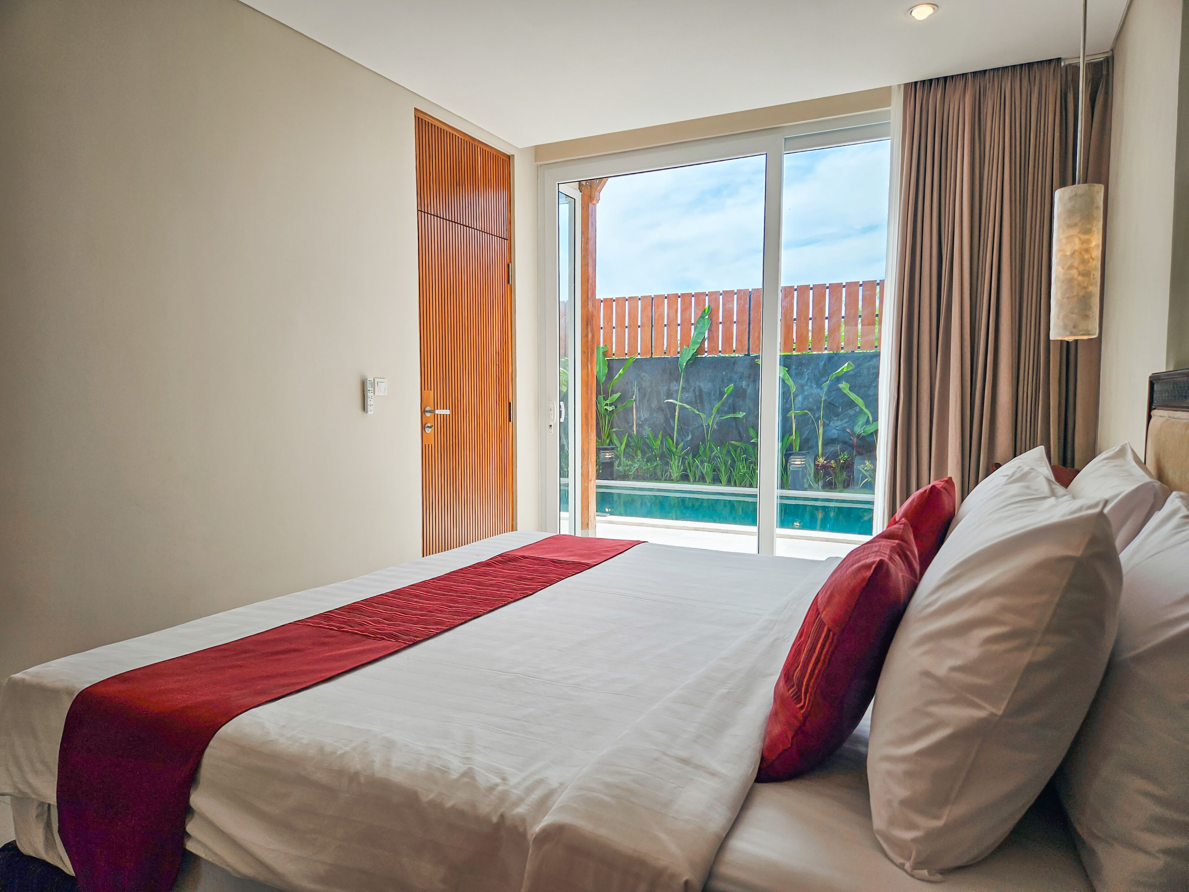 A hotel room with a large bed, a red throw, and a view of a swimming pool and greenery through a sliding glass door.