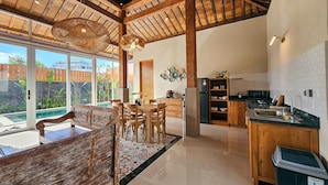 A spacious kitchen with wooden cabinets, a dining area with a large table and chairs, and a view of a swimming pool through glass doors. - Svarna Suite Seminyak (Legian)