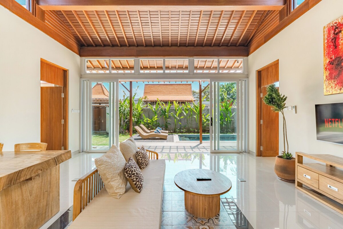 A modern living room with a glass door leading to a pool area, a wooden ceiling, and a TV on the wall.