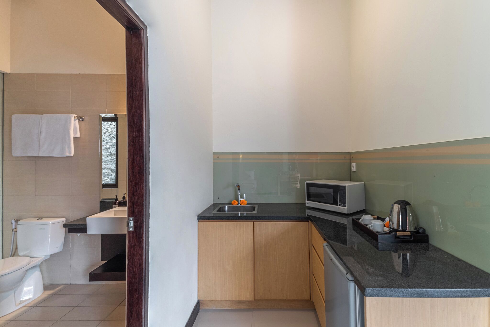 A compact kitchen with a sink, microwave, and kettle, adjacent to a toilet and a small window.