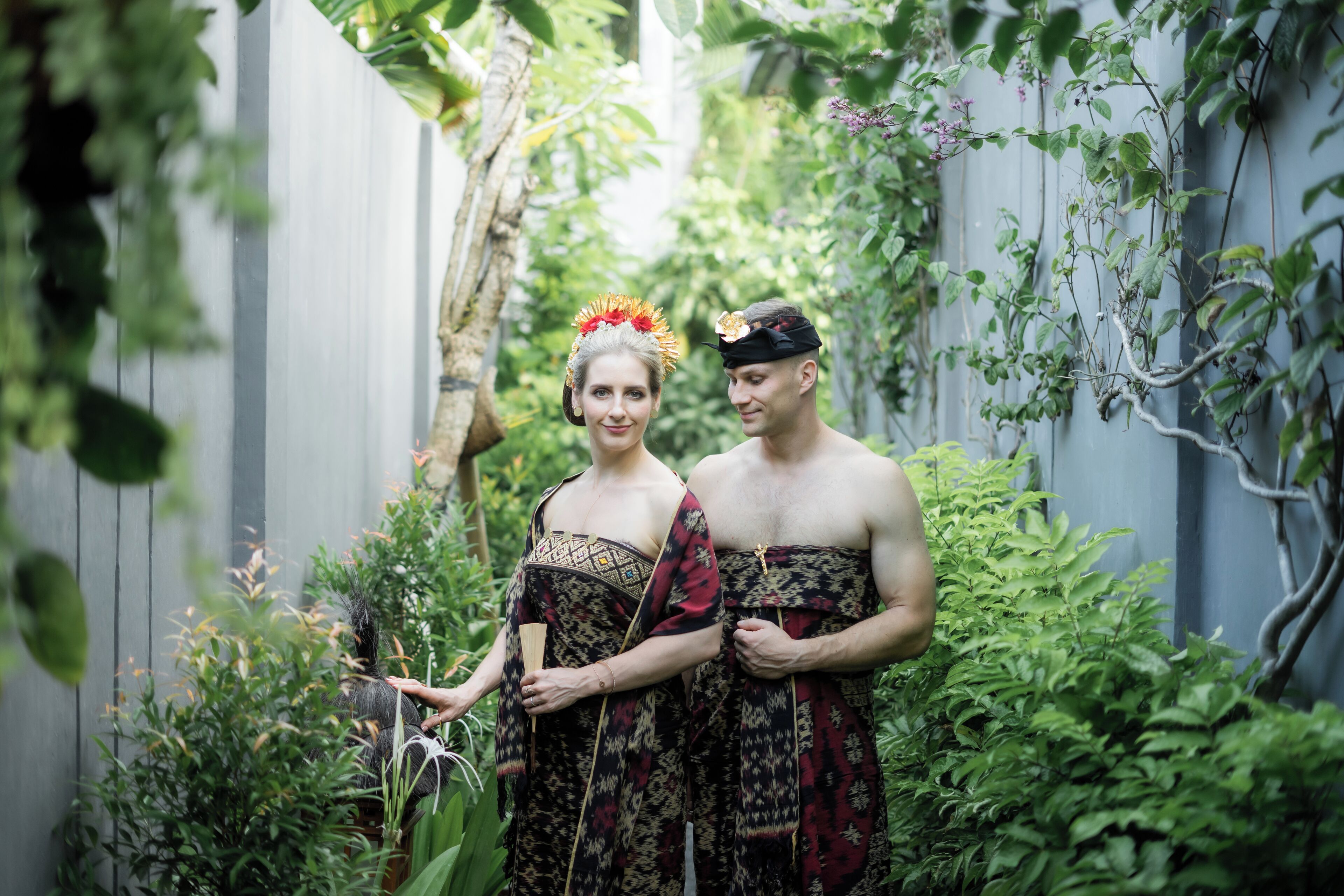 A couple in traditional attire standing in a lush garden with a wooden fence and dense foliage in the background.