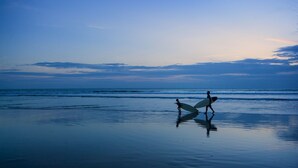 A beach scene with two people carrying surfboards, calm water, and a clear sky. - Samaja Beachside Villas (Seminyak)