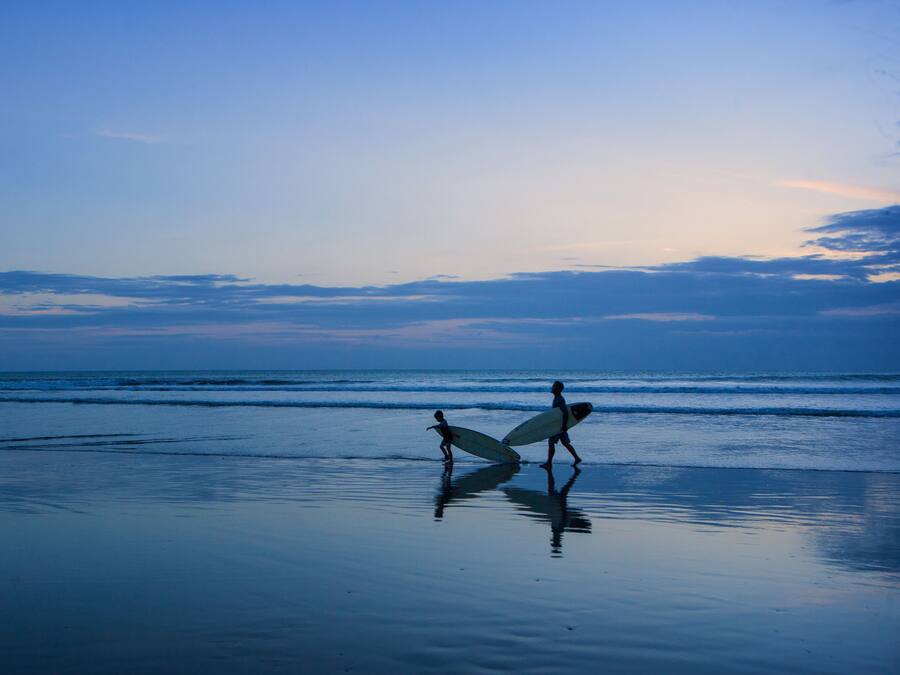 A beach scene with two people carrying surfboards, calm water, and a clear sky.