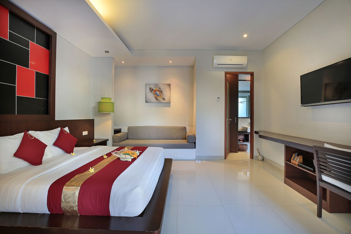 A modern hotel room with a large bed, a sofa, a TV, and a bookshelf.