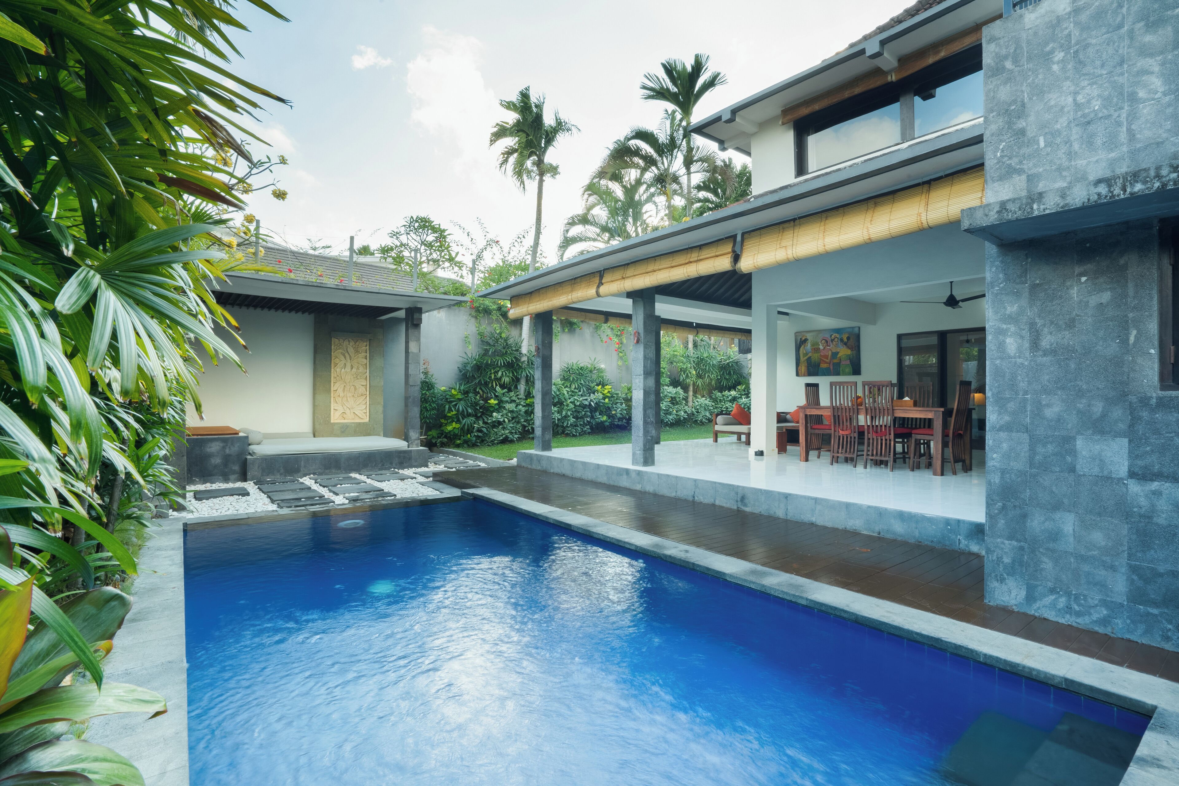 A modern pool area with a clear blue pool, surrounded by lush greenery and a covered seating area with chairs.