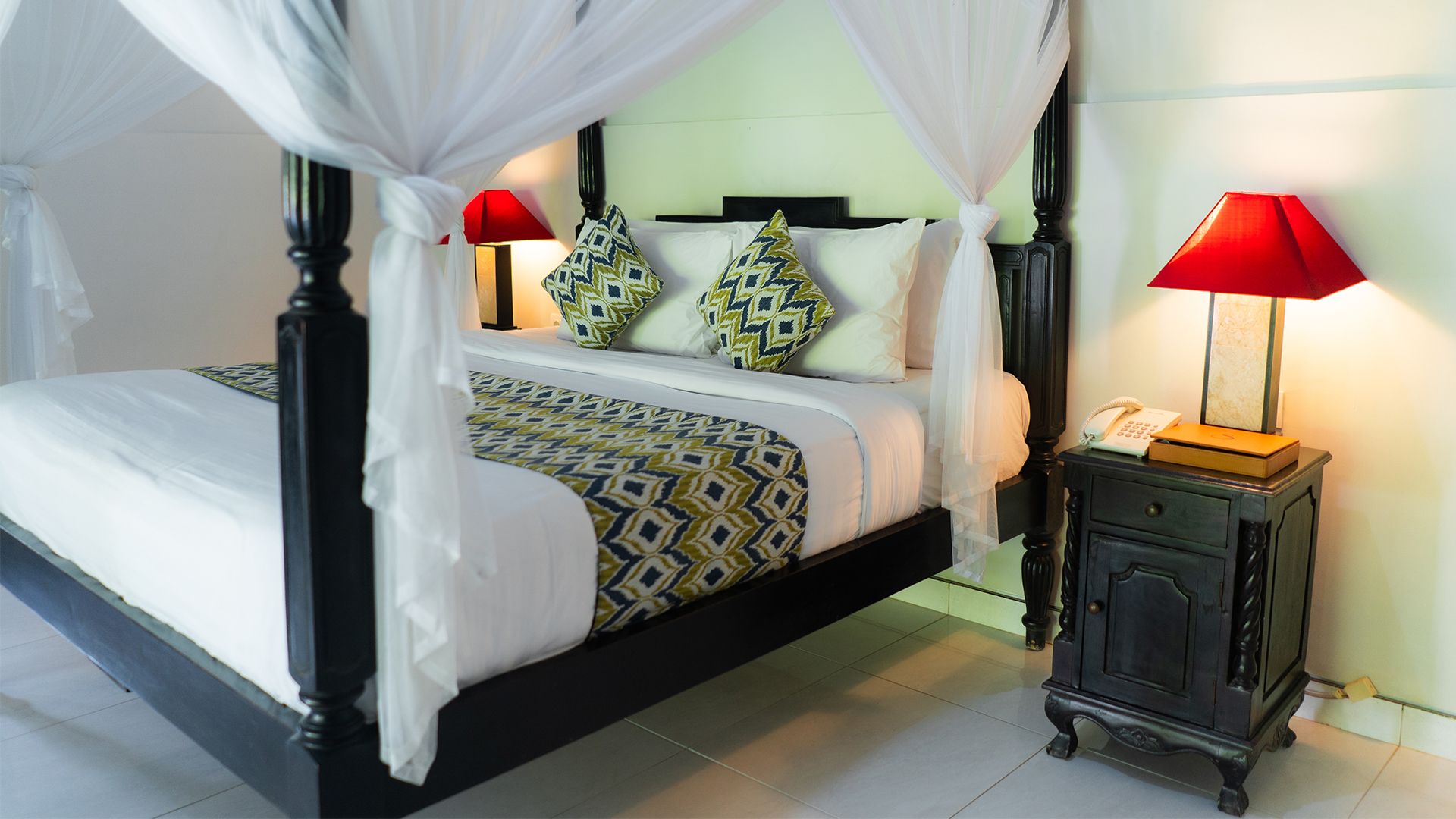 A four-poster canopy bed with patterned bedding, a bedside table with a lamp, and a telephone.