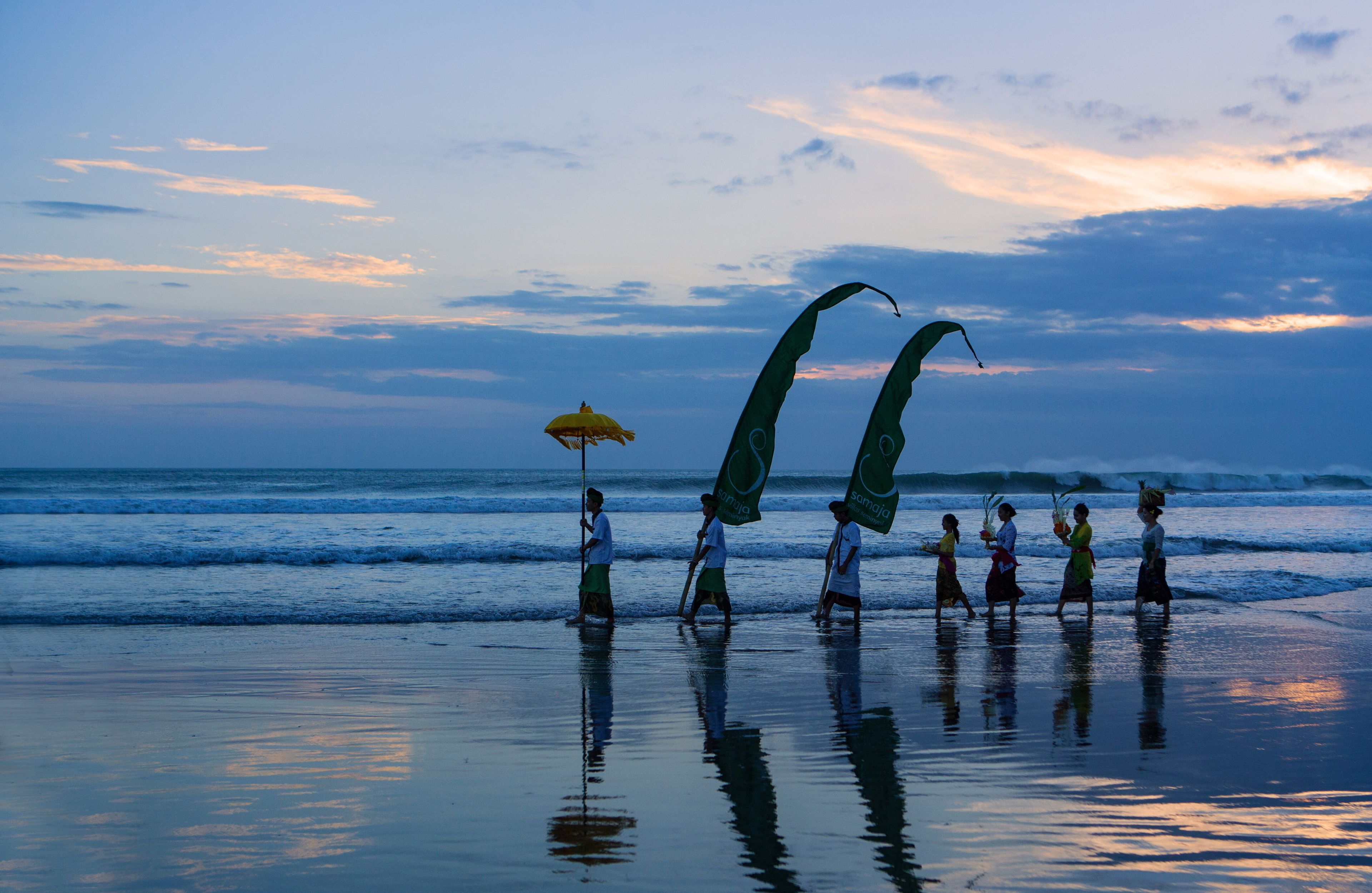 A beach scene with people holding flags and an umbrella, reflecting on the water.