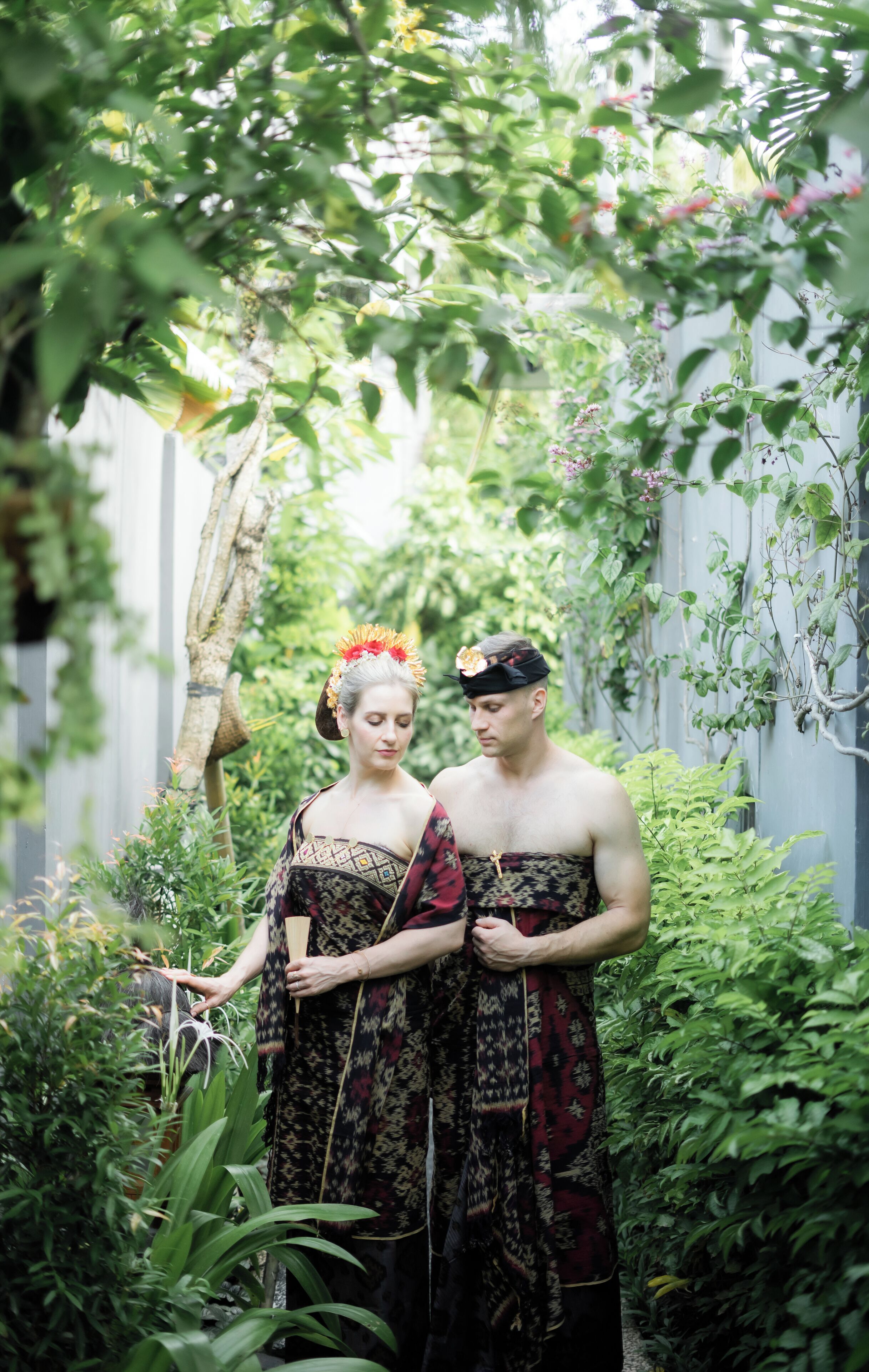 A couple standing in a lush garden with dense foliage and a wooden fence in the background.