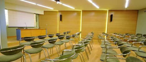 Een vergaderzaal met rijen groene stoelen, een spreekgestoelte, een beamer en een whiteboard.