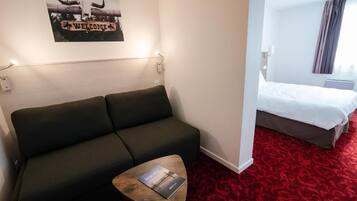 A hotel room with a dark green sofa, a small wooden table with a brochure, a bed with white bedding, and a red patterned carpet.