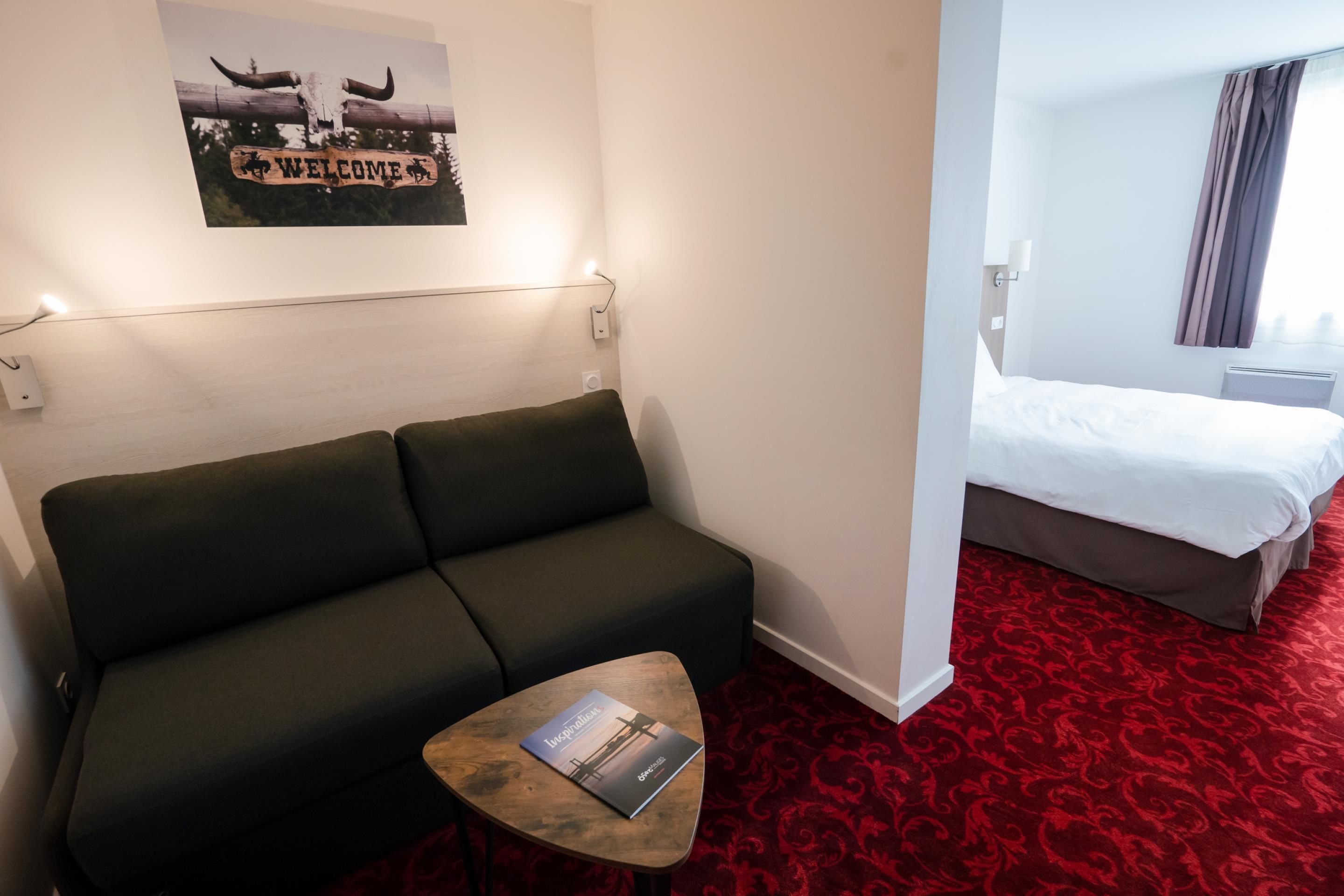 A hotel room with a dark green sofa, a small wooden table with a magazine, a red patterned carpet, a bed with white bedding, and a welcome sign with a cow skull.