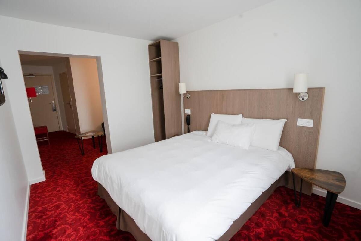 A hotel room with a large bed, a wooden headboard, two bedside lamps, a red carpet, a wooden bench, and a closet with shelves.