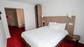 A hotel room with a large bed, a wooden headboard, two bedside lamps, a red carpet, a wooden bench, and a closet with shelves.