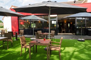 Un espace extérieur aménagé avec des meubles en osier et des parasols, un bar en arrière-plan et une pelouse verdoyante.