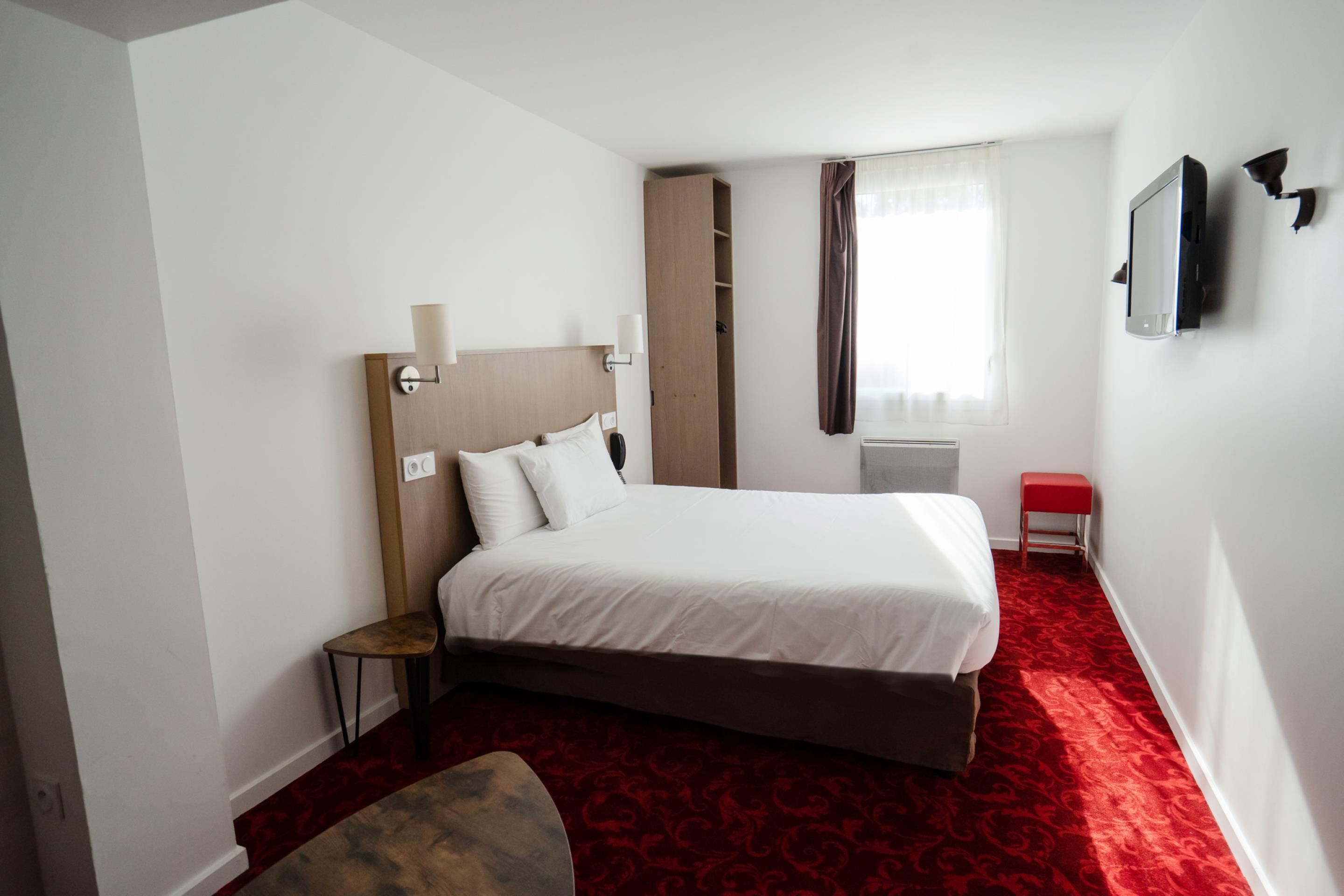 A hotel room with a bed, a small table, a chair, a wardrobe, a TV, and a window with curtains.