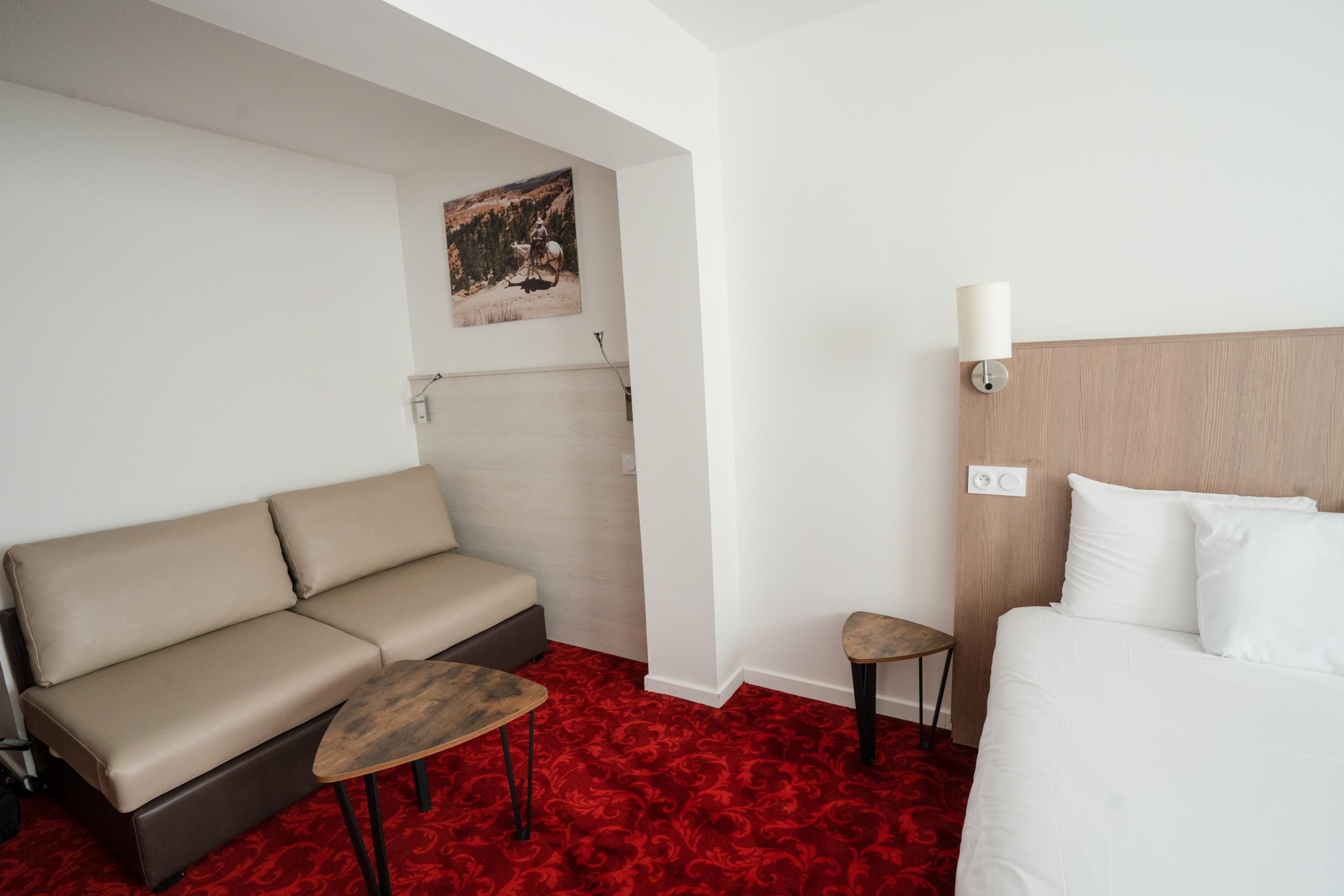 A hotel room with a bed, a sofa, a small table, and a wall-mounted lamp.