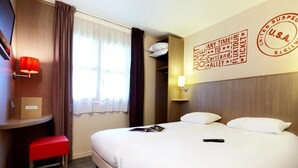 Habitación de hotel con dos camas, un escritorio, un banco rojo, un televisor montado en la pared y una ventana con cortinas.