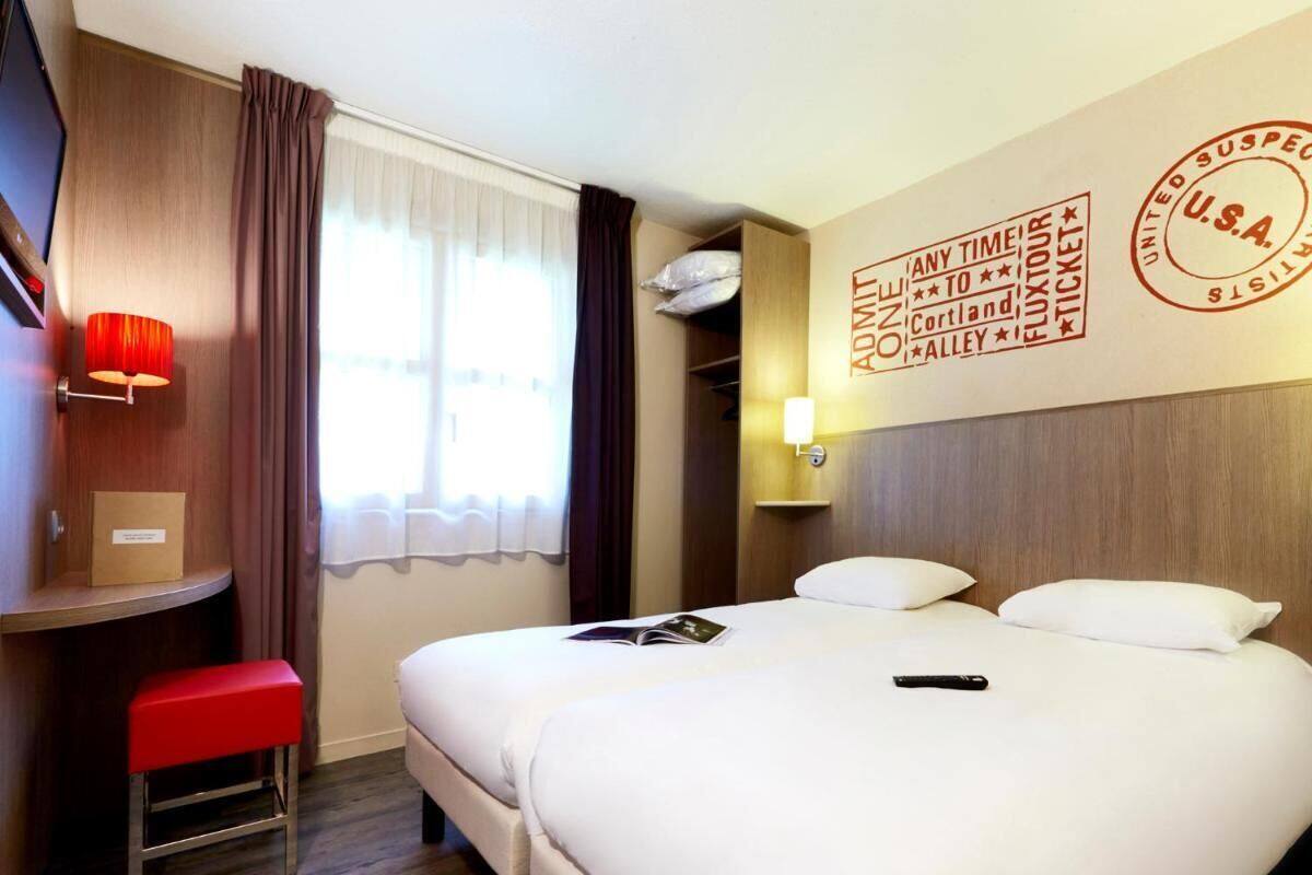 A hotel room with two beds, a desk, a red bench, a wall-mounted TV, and a window with curtains.