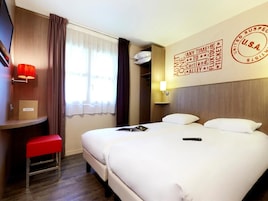 Une chambre d’hôtel avec deux lits, un bureau, un banc rouge, un téléviseur fixé au mur et une fenêtre avec des rideaux.