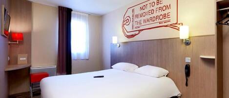 A hotel room with a large bed, a bedside table, a red chair, a wardrobe, and a wall-mounted sign.