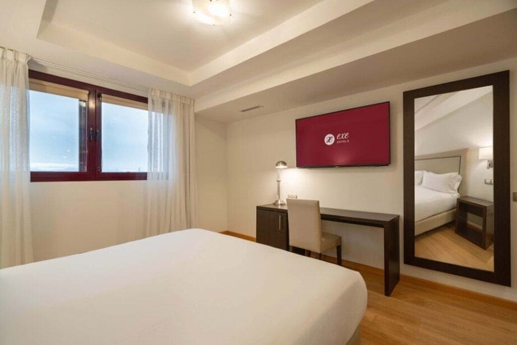A hotel room with a bed, a desk, a chair, a TV, and a large mirror.