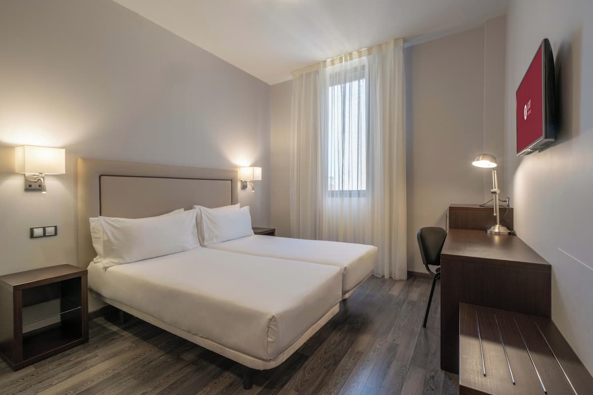 A hotel room with a double bed, bedside lamps, a desk with a chair, a TV mounted on the wall, and a window with curtains.
