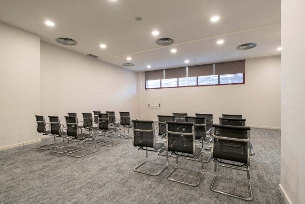 Un salón de conferencias con filas de sillas negras, suelo alfombrado, paredes blancas y una ventana con persianas.