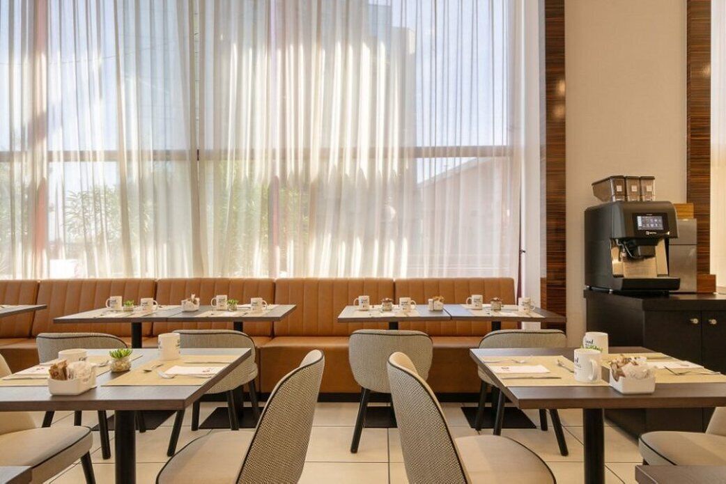 Een modern café met tafels en stoelen, een koffieapparaat en grote ramen met transparante gordijnen.