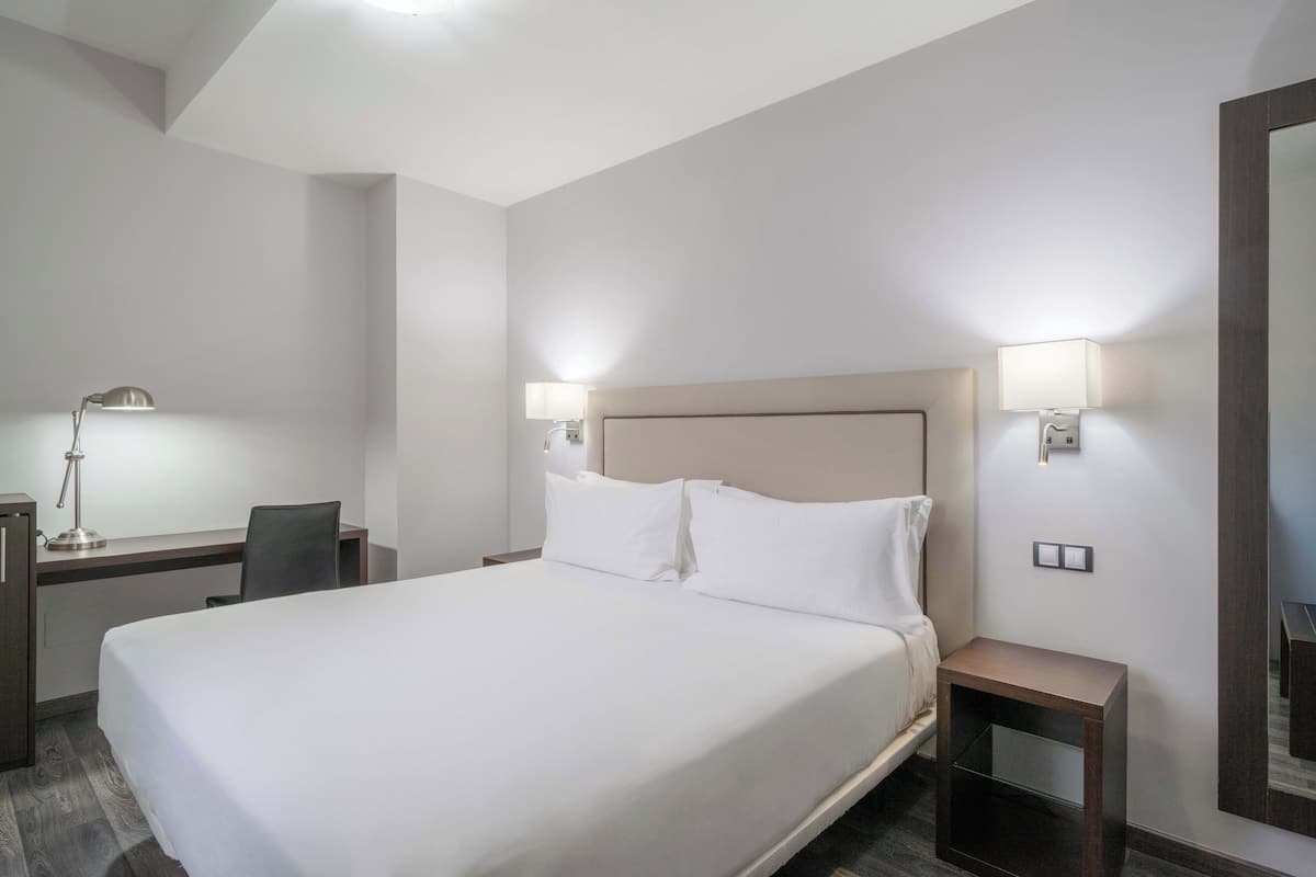 A hotel room with a large bed, a desk with a lamp, and two bedside tables with lamps.