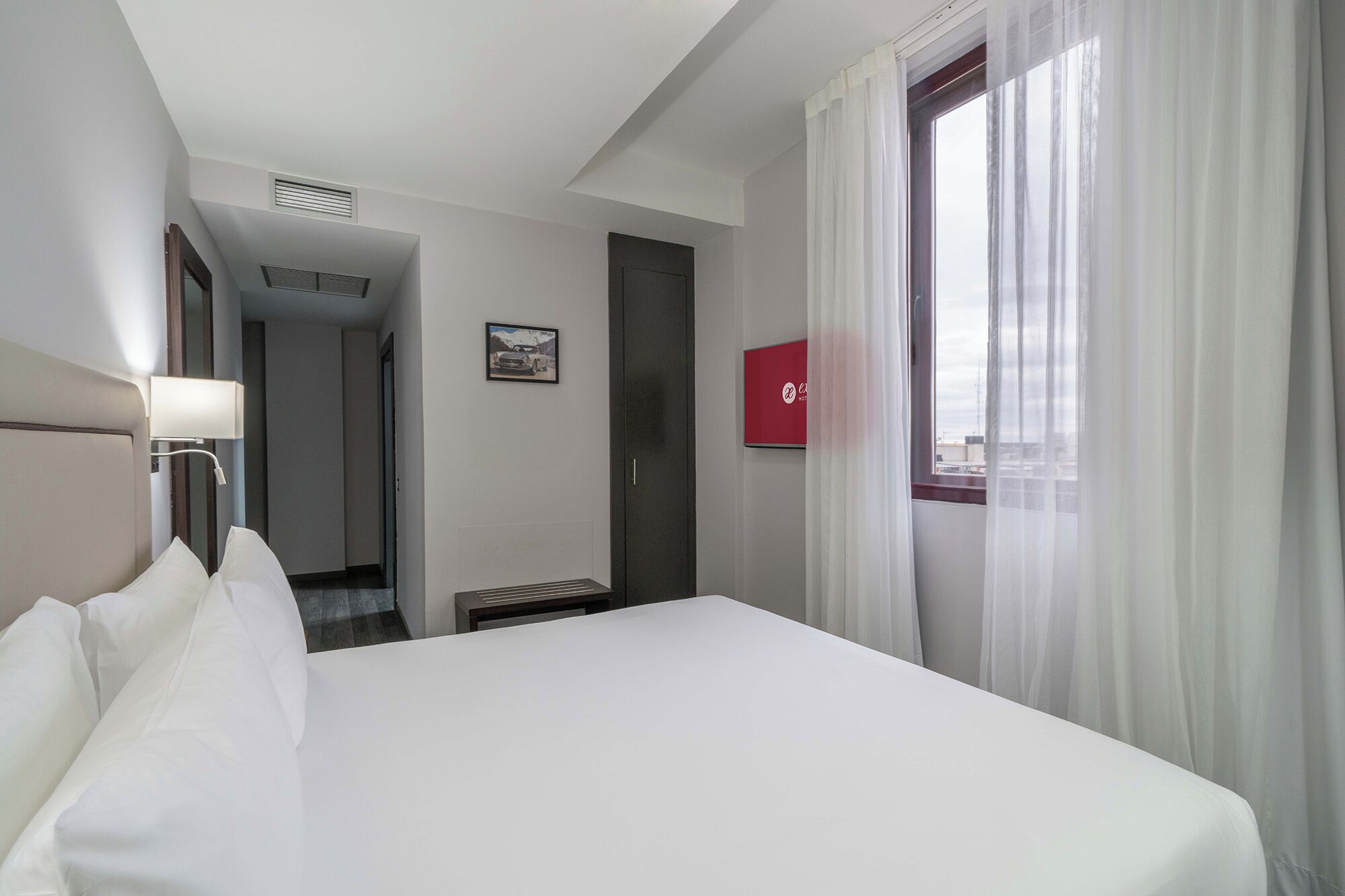 Una habitación de hotel con cama, mesita de noche, lámpara, ventana con cortinas, puerta y pasillo.