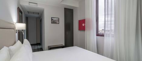 Una habitación de hotel con cama, mesita de noche, lámpara, ventana con cortinas, puerta y pasillo.