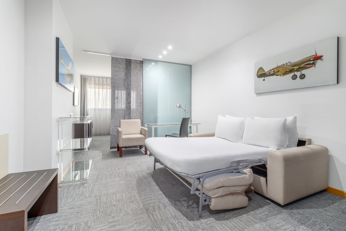 A modern hotel room with a large bed, a desk, a chair, and a glass partition separating the bedroom from a bathroom.