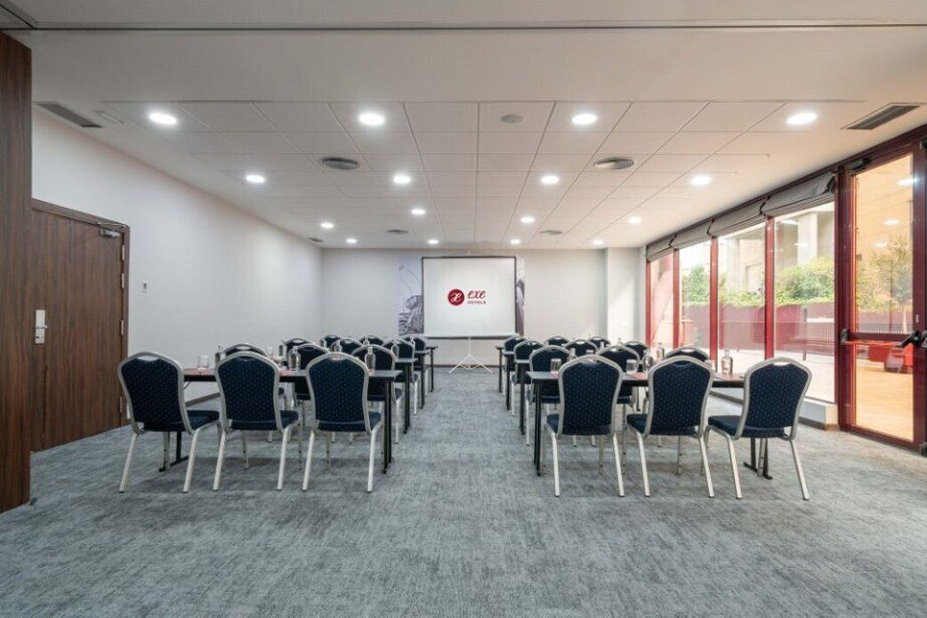Una sala de conferències amb files de cadires orientades cap a una pantalla de projecció, sòl amb moqueta i grans finestres a un costat.