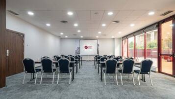 Una sala de conferències amb files de cadires orientades cap a una pantalla de projecció, sòl amb moqueta i grans finestres a un costat.