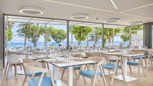Un restaurante moderno con mesas y sillas dispuestas para comer, ventanas amplias con vistas al mar y árboles, y un techo con lámparas circulares.