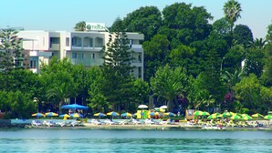 Un hotel con un letrero que dice "Hotel Triton" en un edificio con balcones, rodeado de exuberante vegetación y una playa con sombrillas de colores.