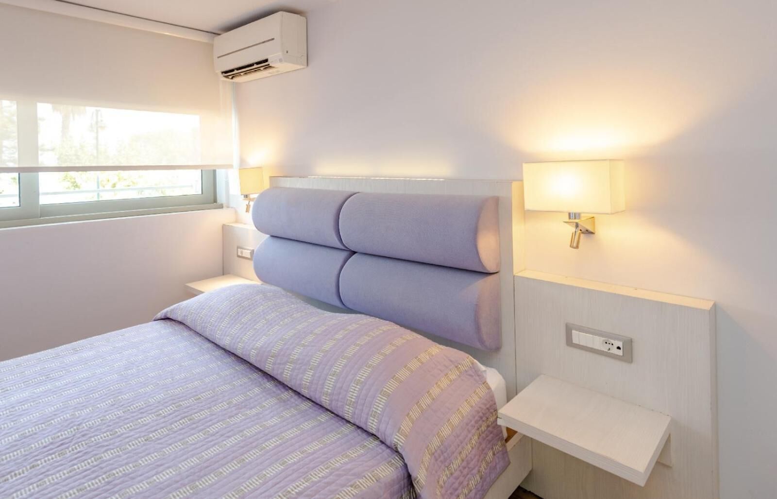 A hotel room with a bed, bedside tables, a wall-mounted air conditioner, and a window with a view.