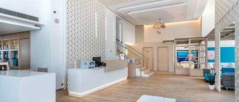 A spacious, modern hotel lobby with a reception desk, a staircase, and a seating area with a view of the sea.