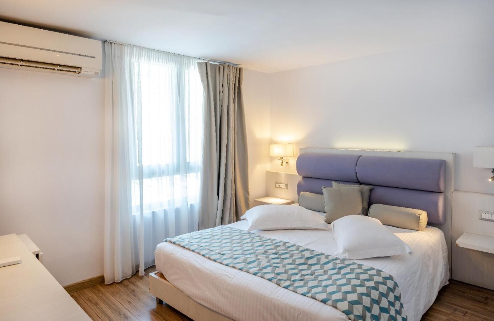 Een hotelkamer met een bed, nachtkastje, lamp en raam met gordijnen.