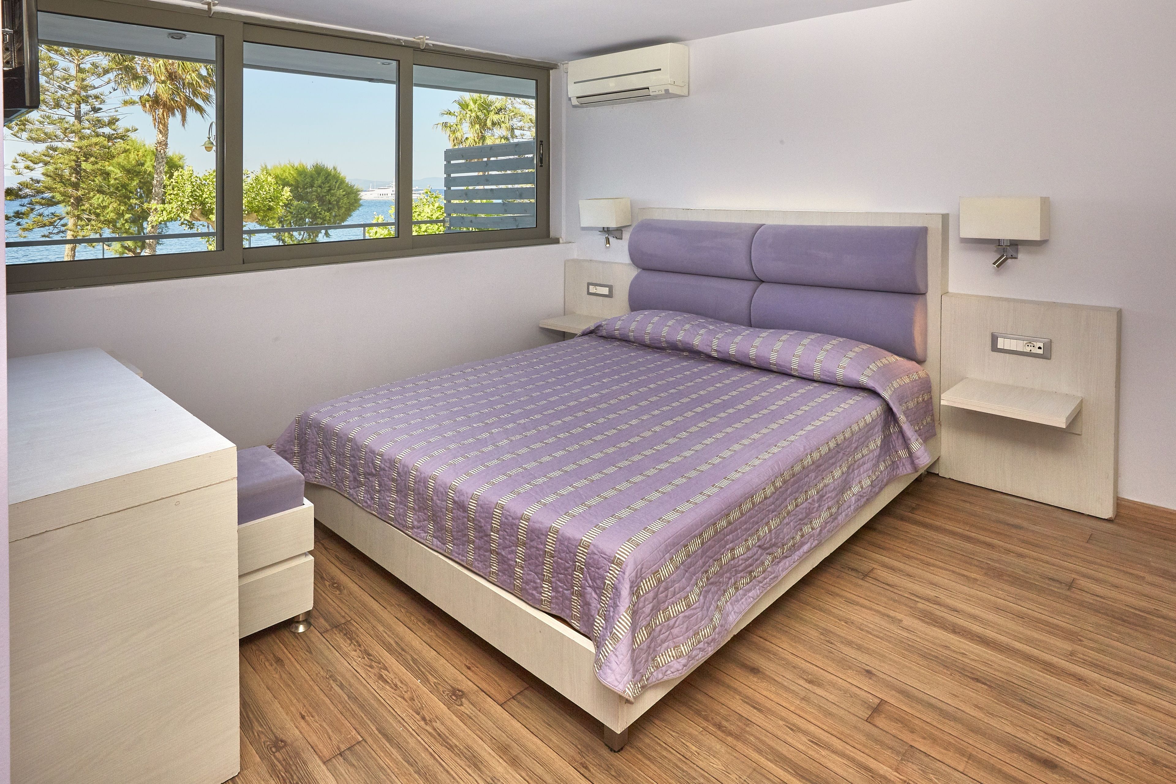 Un dormitorio con cama, mesitas de noche, escritorio y ventana con vistas a árboles y un cuerpo de agua.
