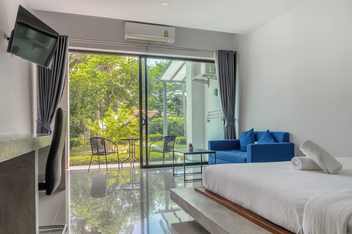 A modern hotel room with a large sliding glass door leading to a balcony, a bed with white bedding, a blue sofa with pillows, and a flat-screen TV mounted on the wall.