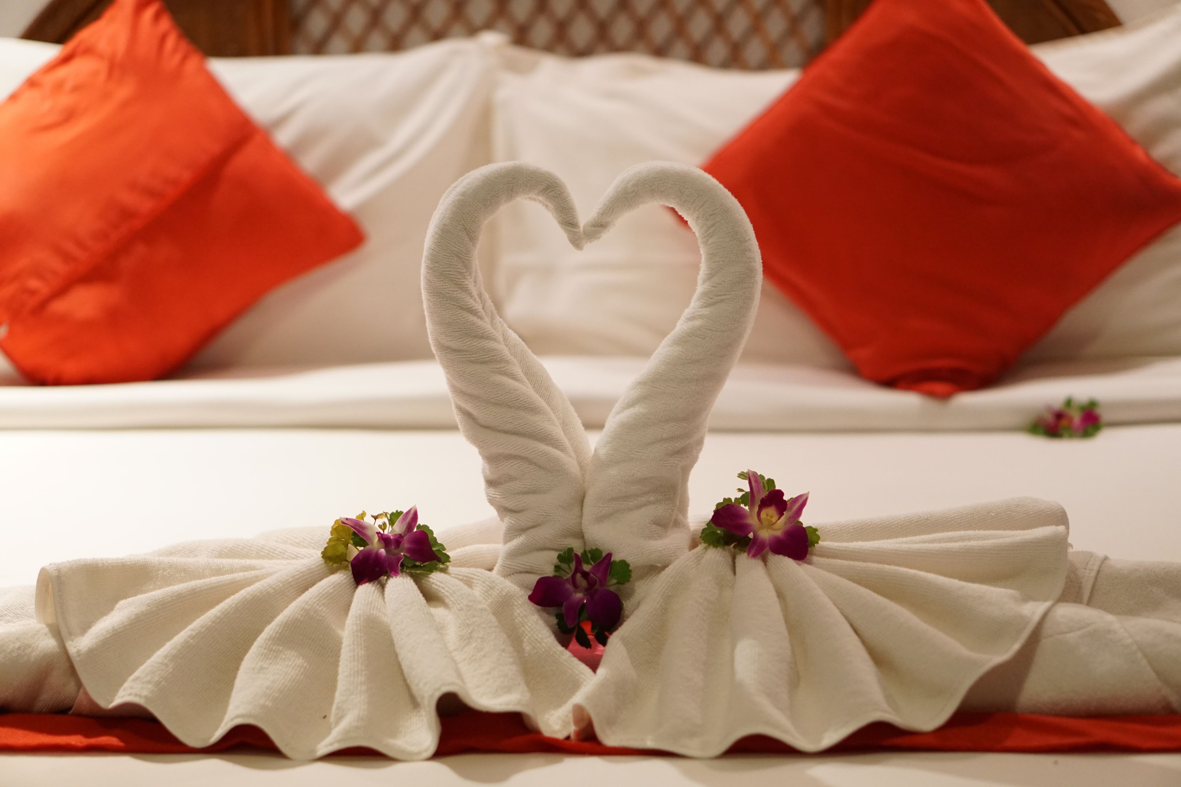 A bed with white linens and red pillows, a heart-shaped towel arrangement, and purple flowers.
