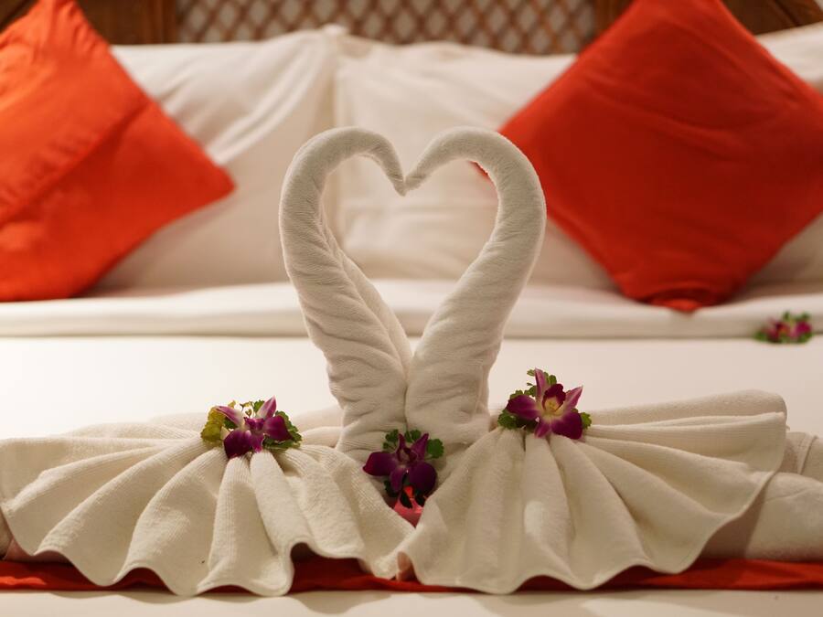 A bed with white linens and red pillows, a heart-shaped towel arrangement, and purple flowers.