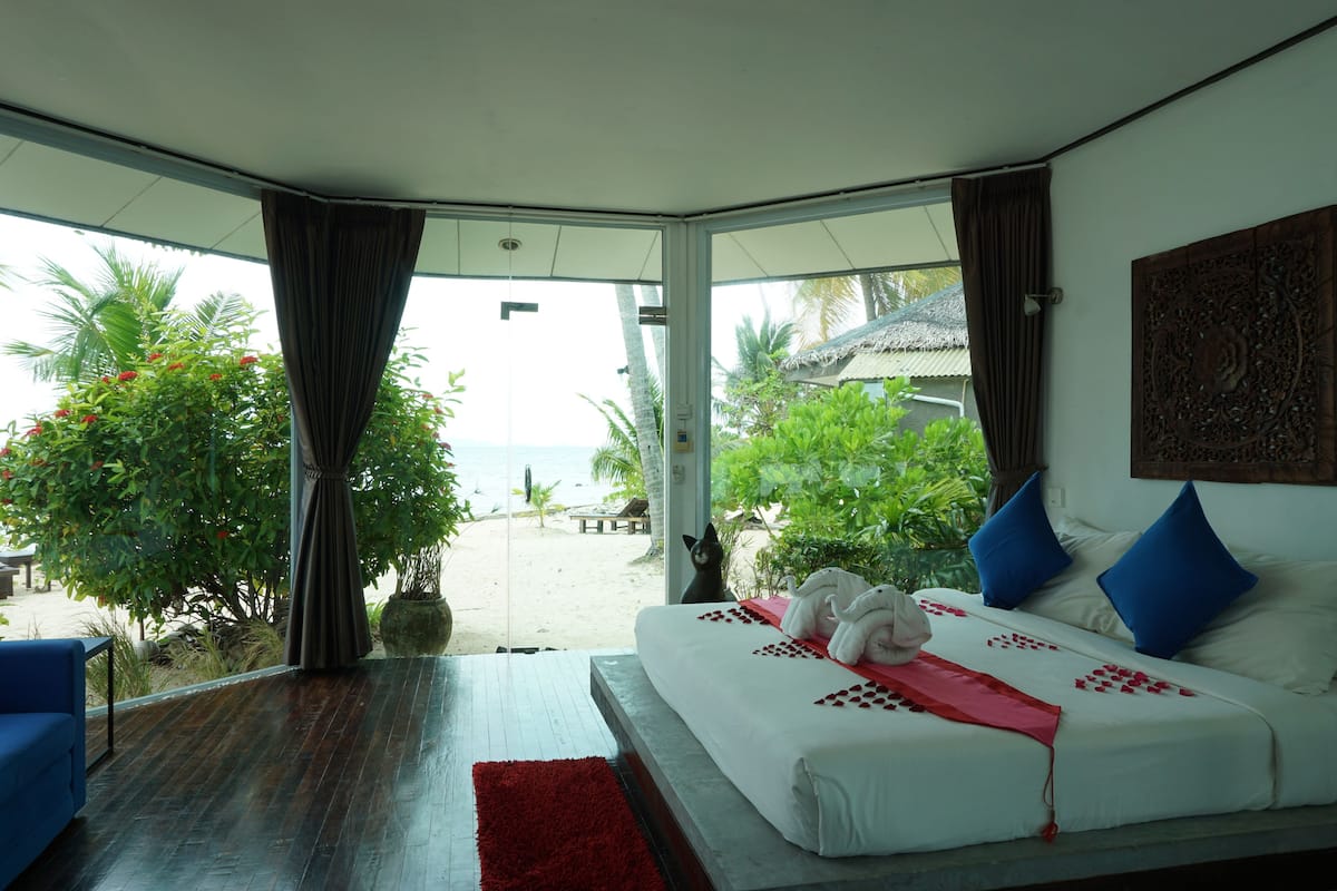 A bedroom with a large window, a bed with white linens, blue pillows, and a red throw. There is a blue sofa and a red rug on the floor.